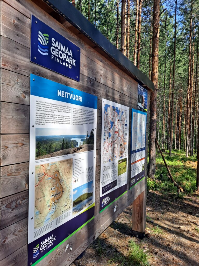 Puinen seinä, jossa ylhäällä sininen kyltti, jossa lukee Saimaa Geopark Finland. Alalaidassa kolme julkistetta, joissa vasemmmanpuoleisessa lukee ylhäällä Neitvuori. 